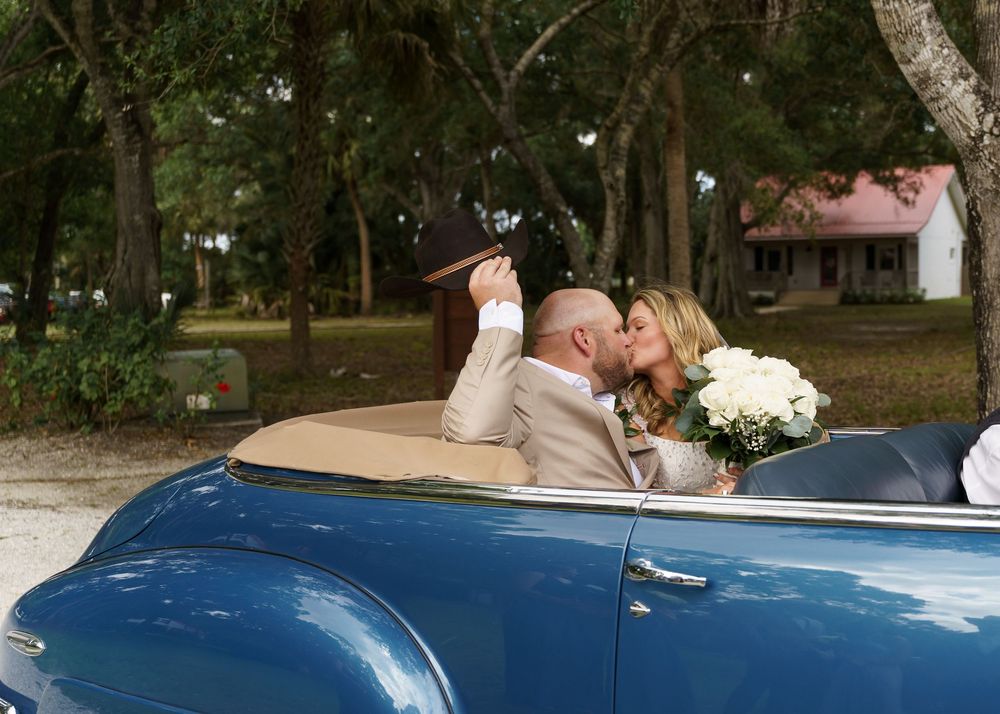 Newlywed couple kissing in a vintage blue convertible car, bride holding white bouquet, groom in tan suit and hat.