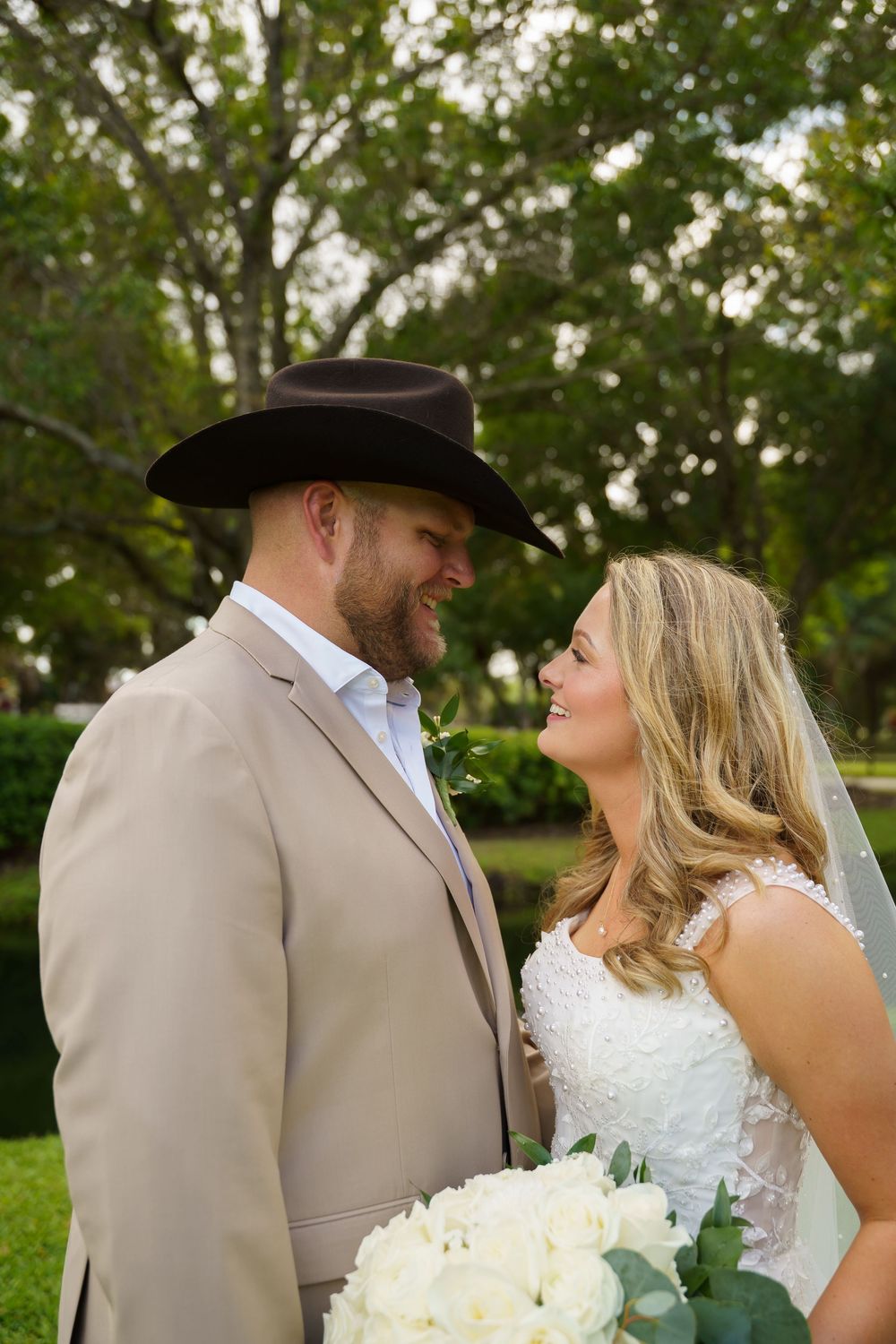Smiling bride and groom in tan suit and black cowboy hat share a romantic moment outdoors among green trees.