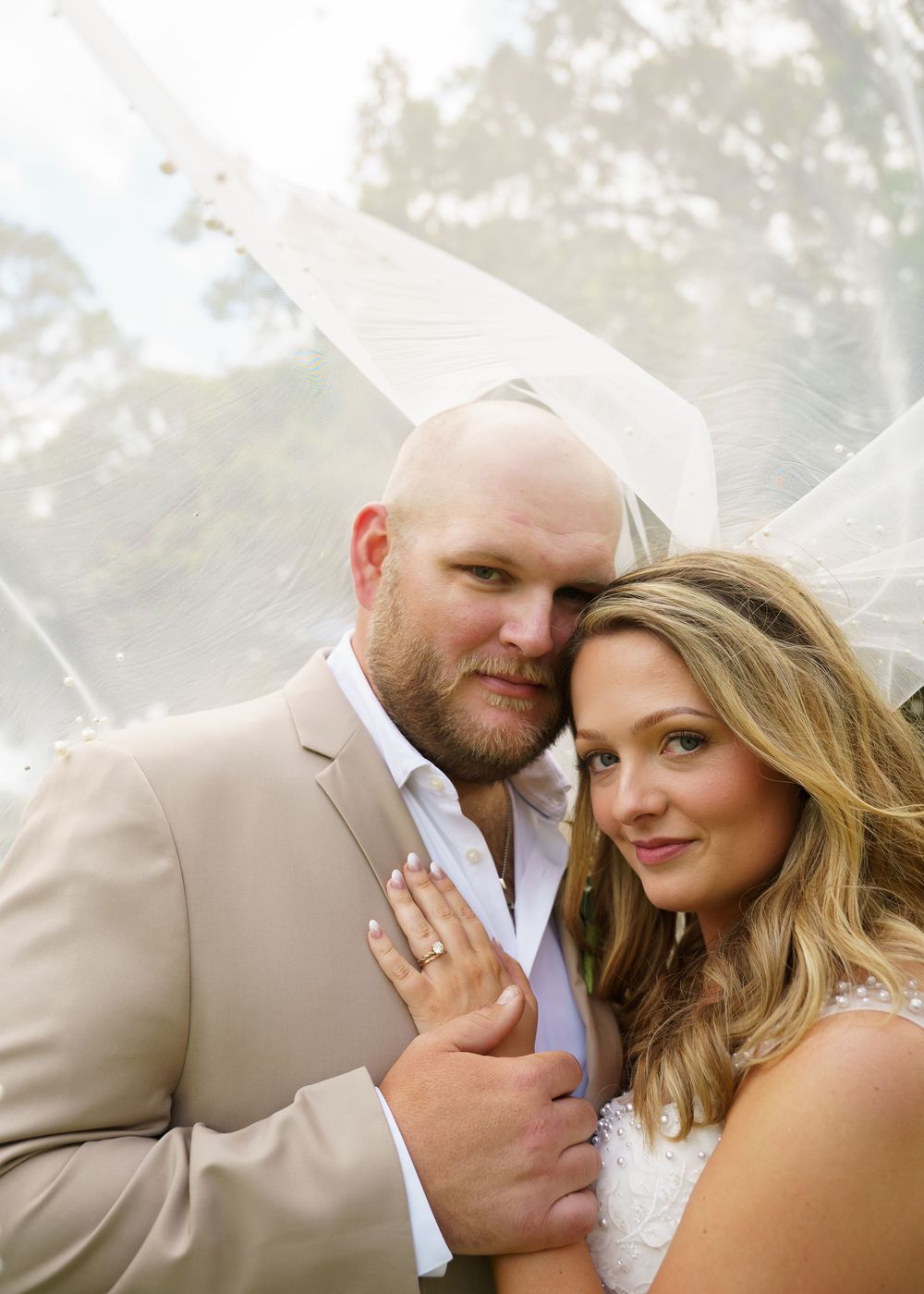 Bride and groom pose romantically under a veil in an outdoor wedding portrait.