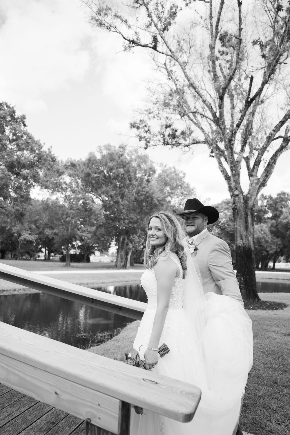 Black and white photo of a bride and groom posing on a wooden bridge near a pond surrounded by trees.