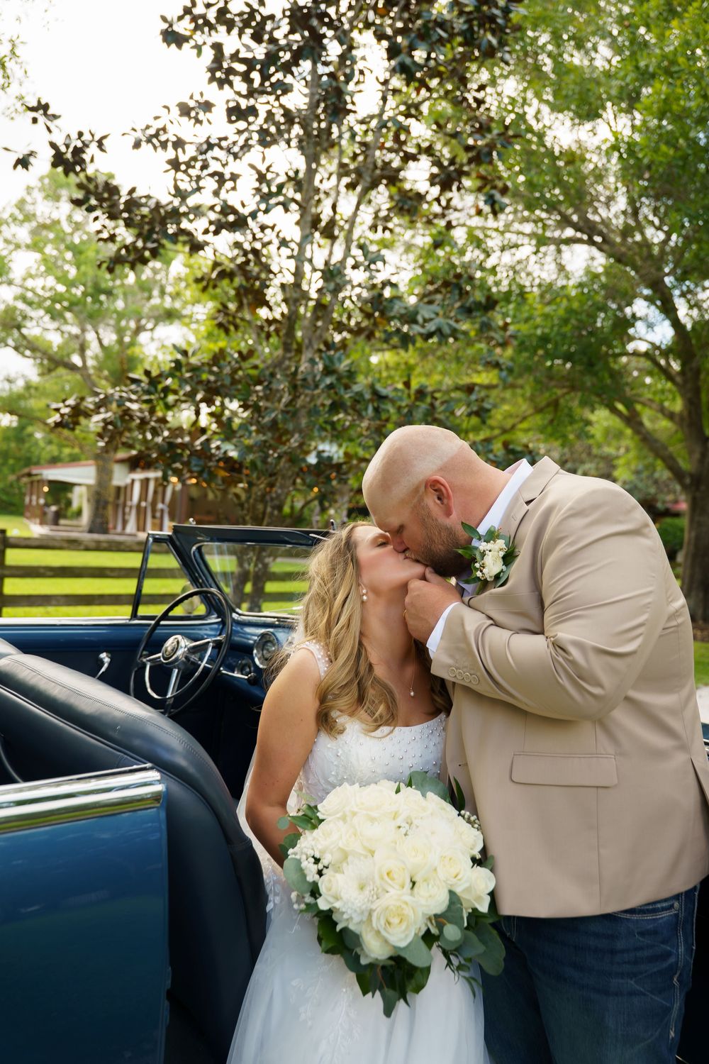 Bride and groom share a romantic kiss beside a vintage blue convertible car, surrounded by lush green trees.