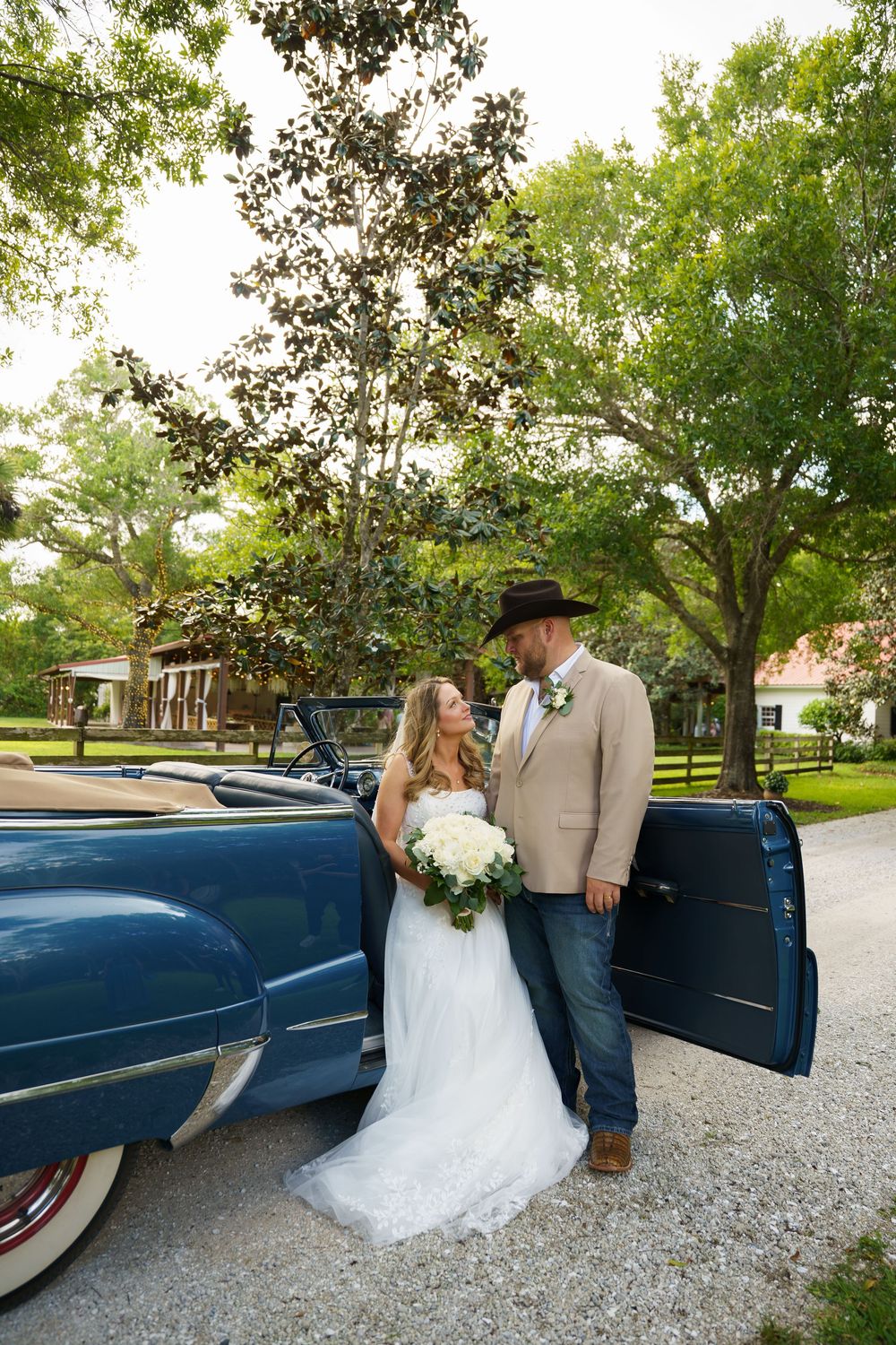 Bride and groom pose beside a vintage blue convertible car surrounded by lush green trees on their wedding day.