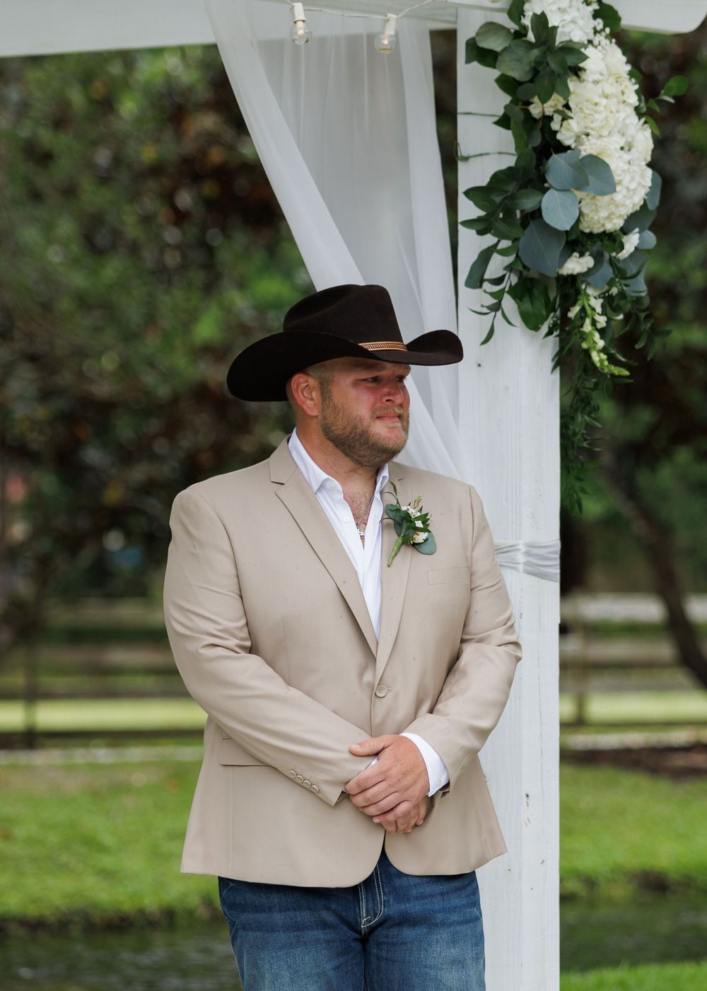 Groom in tan blazer, cowboy hat, and boutonniere stands at outdoor wedding altar with white draping and floral decor.