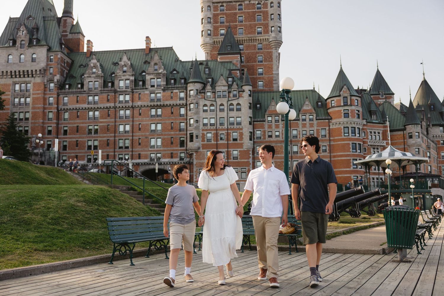 Family of four walking on a boardwalk near Château Frontenac in Quebec City on a sunny day.