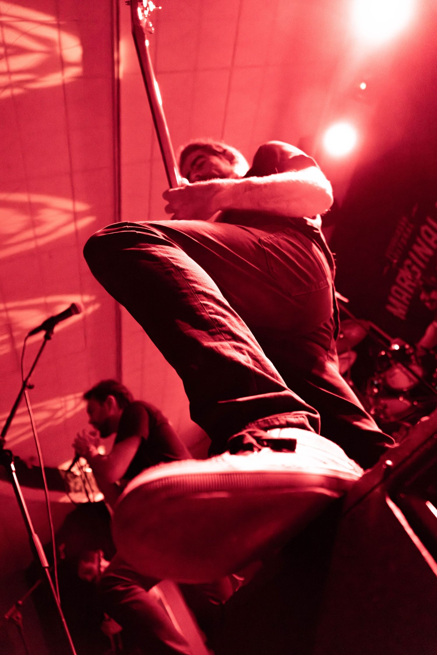A guitarist leaps on stage bathed in red light during an energetic live rock concert performance.