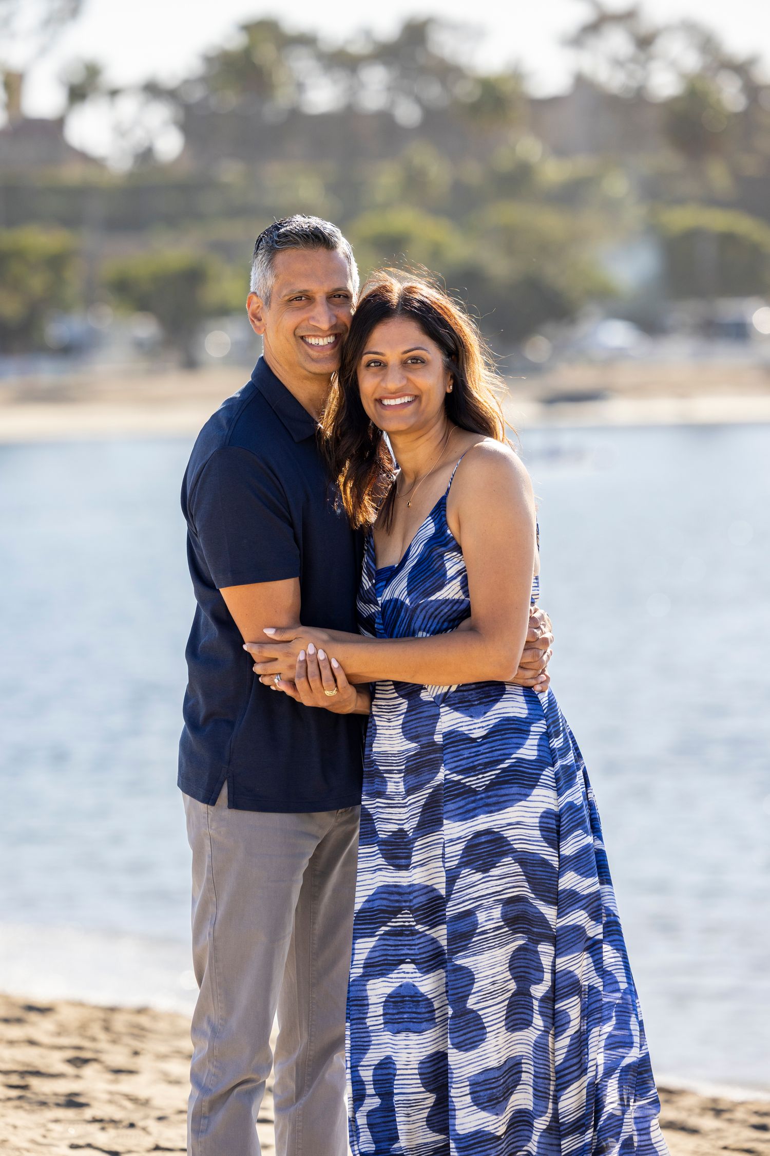 A smiling couple embraces on a sandy beach, woman in blue patterned dress, man in navy shirt, with water in background.