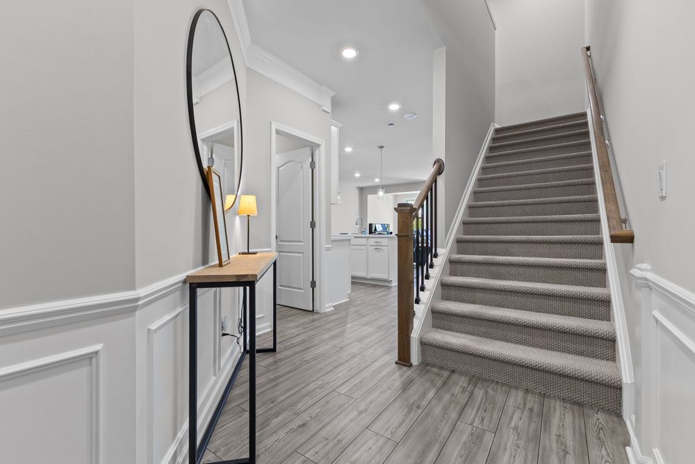Bright home entryway featuring wainscoting, round mirror, staircase with carpet runner, and hardwood-style flooring.