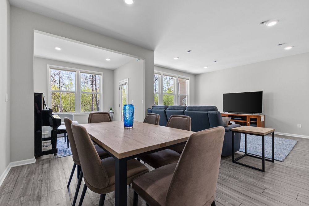 Open-concept dining and living area with wooden table, upholstered chairs, and large windows overlooking trees.
