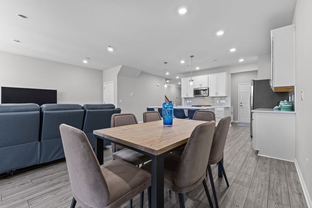 Modern open-plan dining area with wooden table, gray chairs, white kitchen, and recessed lighting throughout.