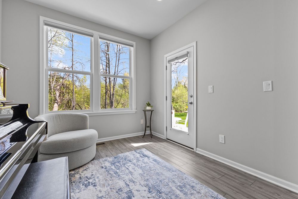 Bright sitting room with large windows, glass door to backyard, gray accent chair, and hardwood-style floors.