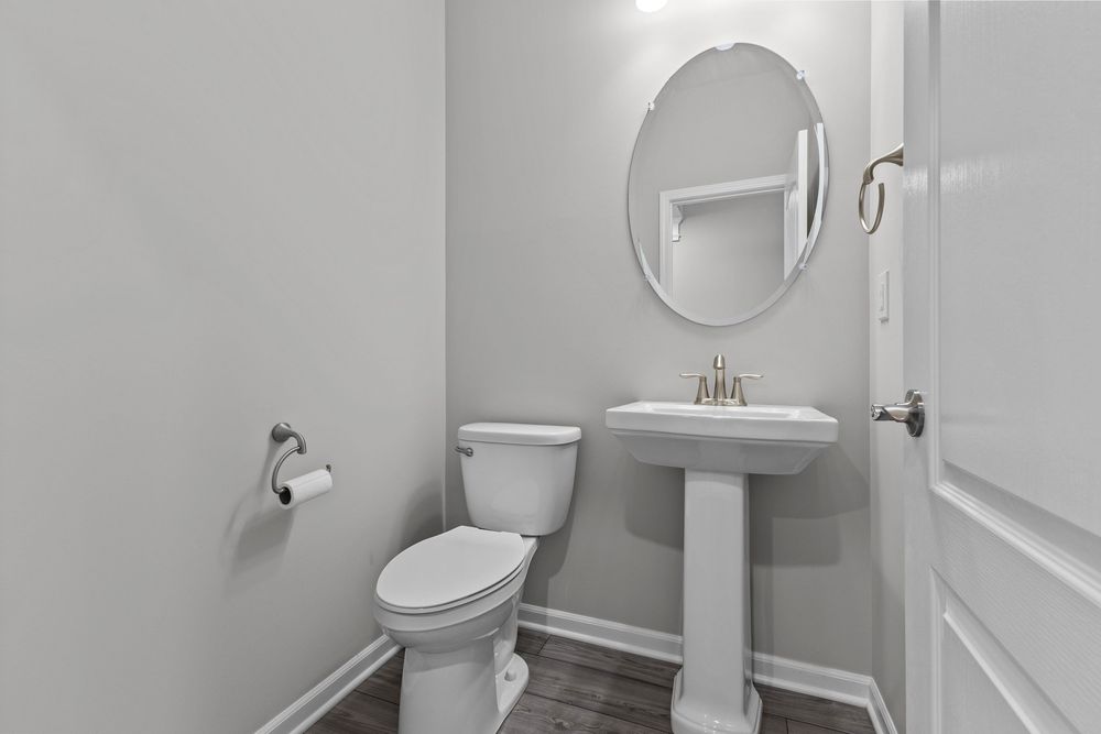 Small powder room with white pedestal sink, round mirror, toilet, and gray walls on wood-look tile floor.