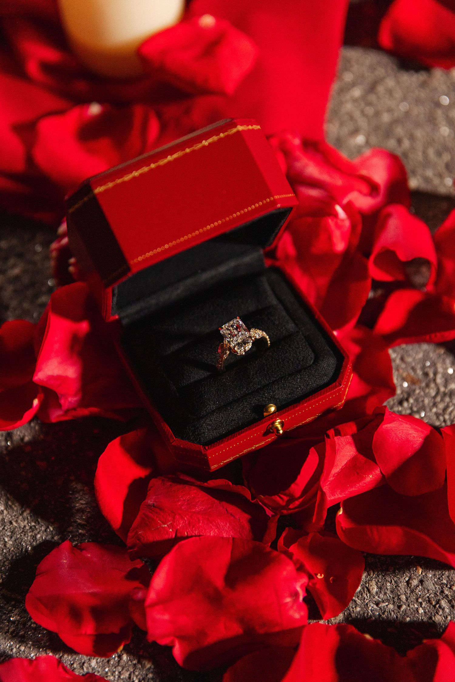 Diamond engagement ring in red Cartier box surrounded by red rose petals and candle light.