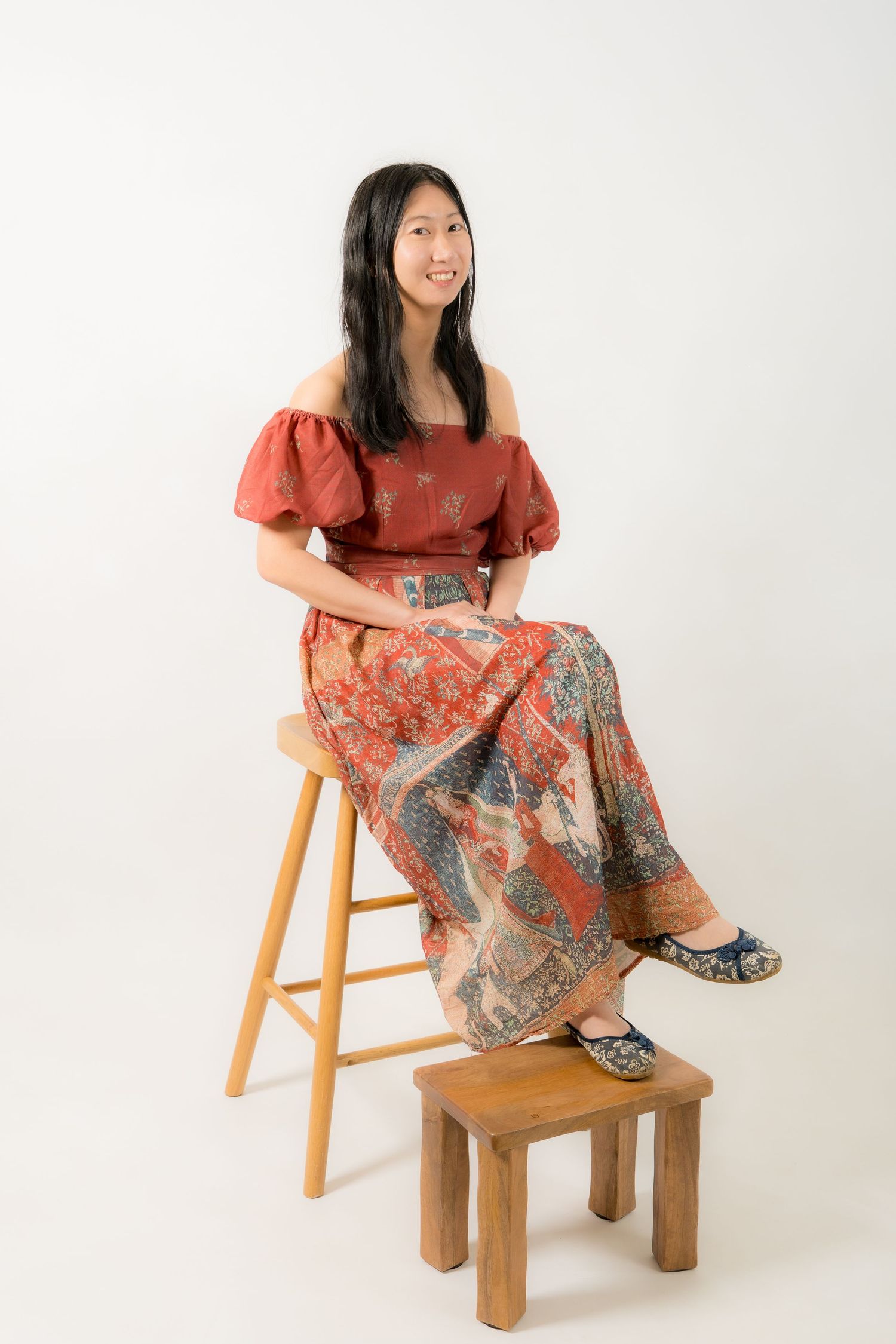 Woman in red patterned off-shoulder dress with puff sleeves and maxi skirt, posing beside white pedestal.