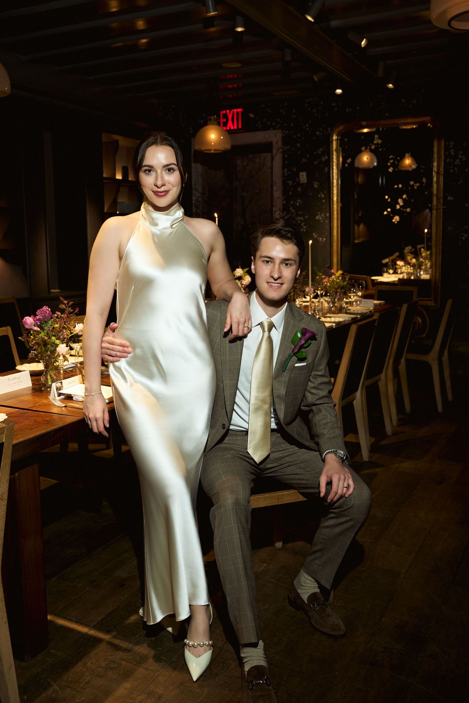 Elegant couple posing in a dimly lit restaurant, bride in white satin gown and groom in gray suit.