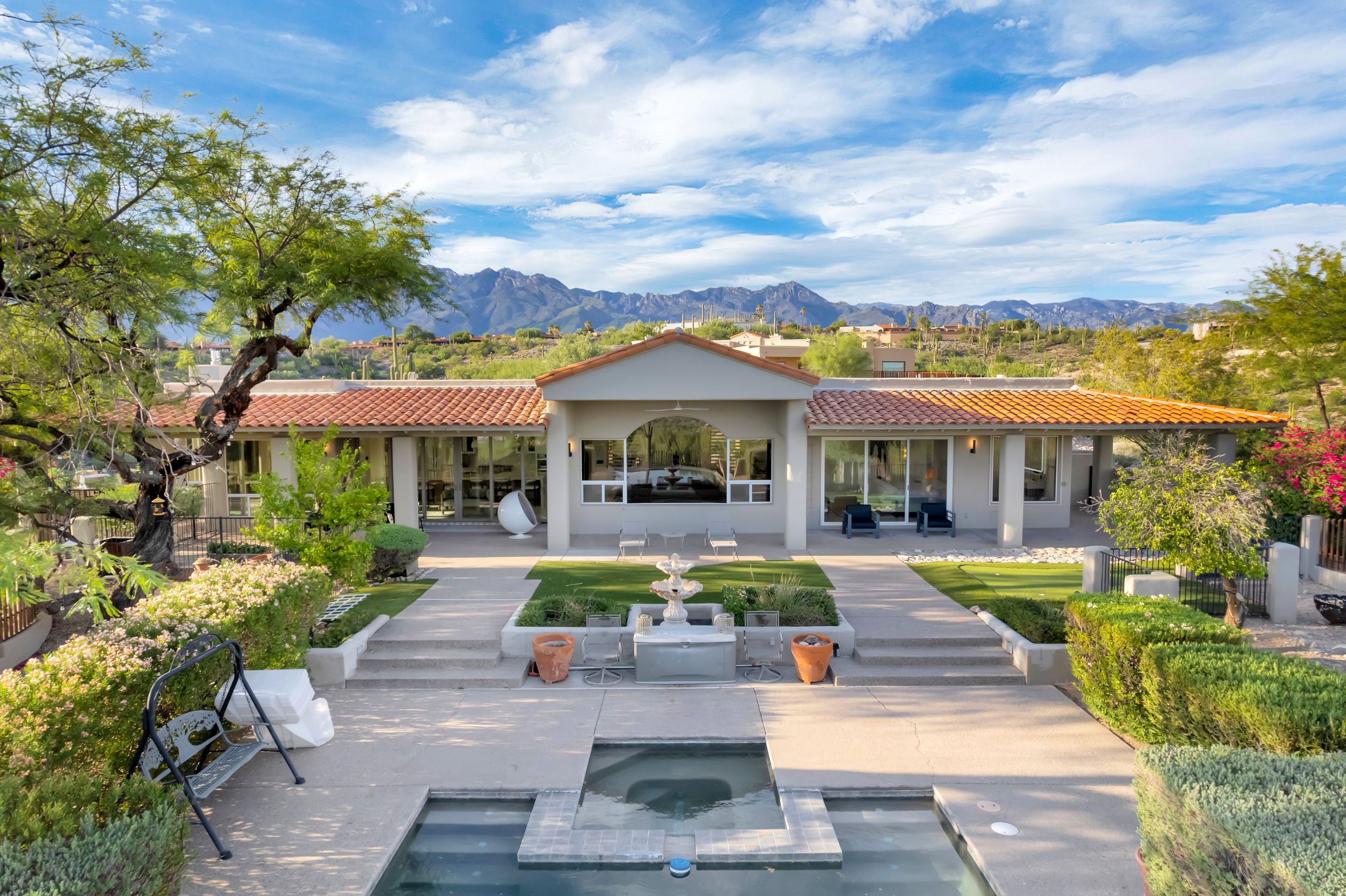Elegant ranch-style home with fountain, pool, lush landscaping, and stunning mountain views under a blue sky.