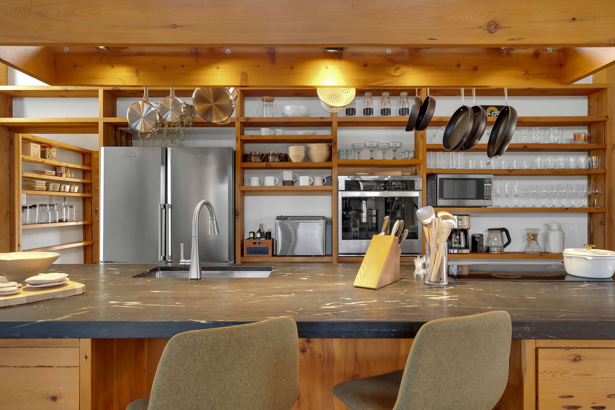 Modern kitchen interior with wood cabinets, stainless steel appliances, and open shelving displaying glassware.