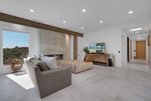 Modern living room with marble floors, gray sectional sofa, wall-mounted TV, and panoramic windows overlooking a hillside.