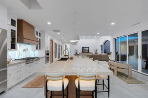 Modern open-concept kitchen with white cabinets, marble island, gold bar stools, and large windows overlooking outdoor living area.