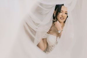 Radiant bride in lace wedding dress smiles through flowing white veil with soft natural light.