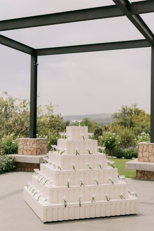 Elegant tiered white wedding cake display with greenery accents, set outdoors under a modern pergola with garden views.