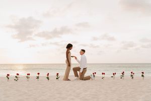 Romantic beach proposal at sunset with rose line on sand, man on one knee offering ring to woman.