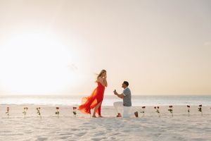 Man proposes on a sandy beach at sunset, woman in red dress, roses lining the shore.