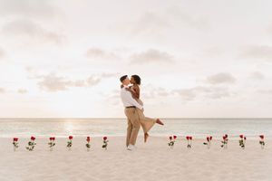 Couple sharing a romantic kiss on a sandy beach at sunset, surrounded by a row of red roses.