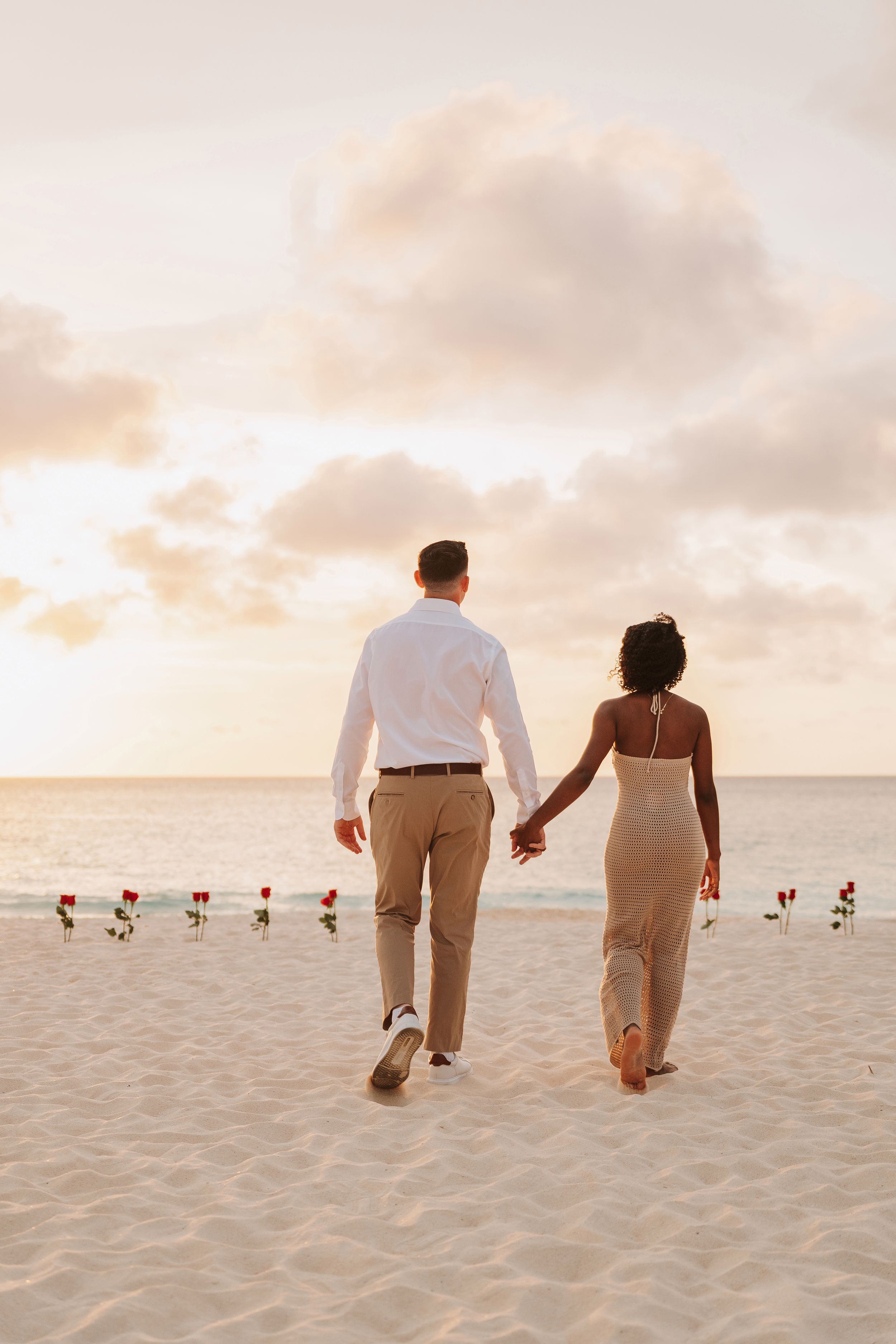 Couple holding hands walking on a romantic beach at sunset with red roses lining the sandy shore.