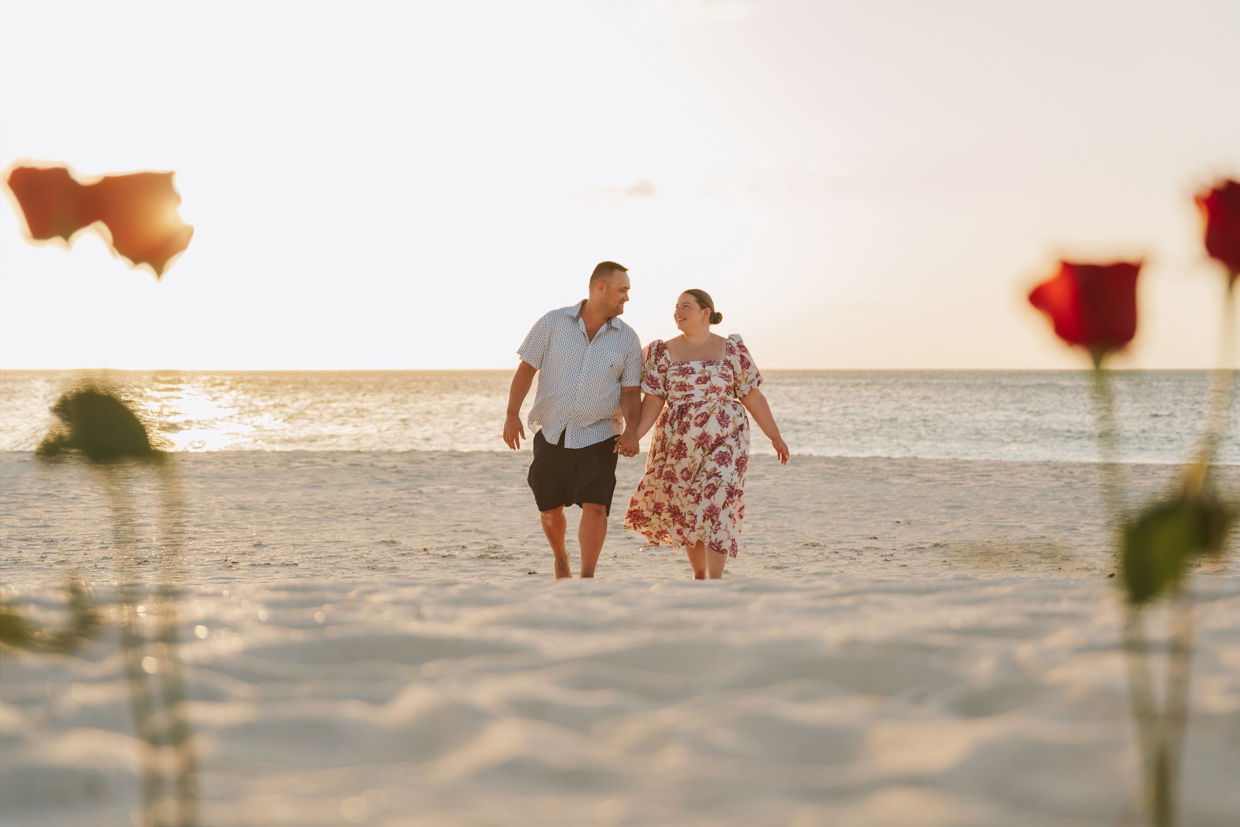 Couple walking hand-in-hand on a sandy beach at sunset, framed by red roses in the foreground.