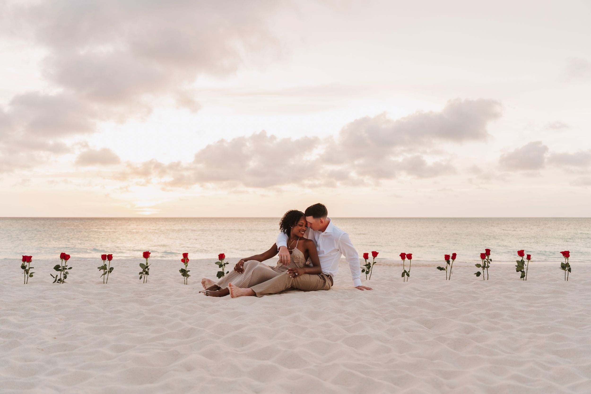 Couple sitting on a sandy beach at sunset surrounded by a row of red roses in a romantic proposal setting.