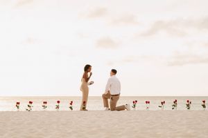 Man proposes on a sandy beach with red roses, woman covers mouth in surprise during romantic sunset proposal.