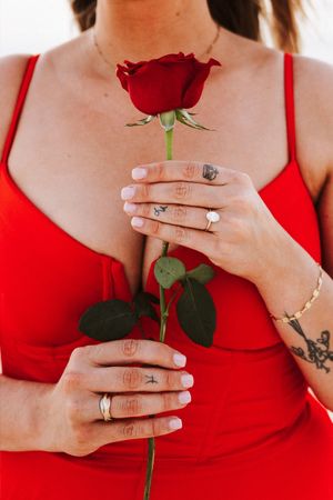 Woman in red dress holding a single red rose, wearing a diamond engagement ring and finger tattoos.