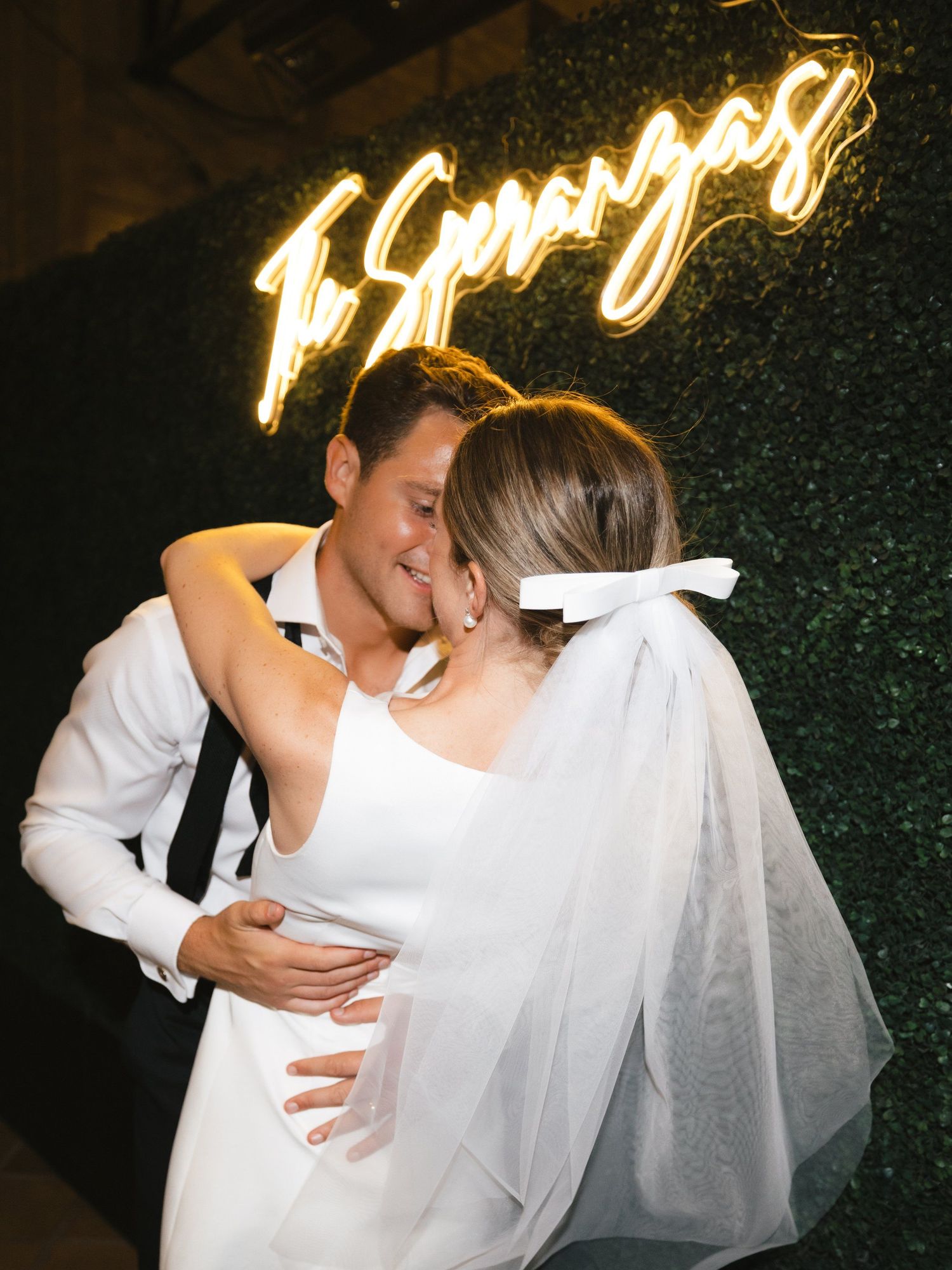 Newlywed couple kissing in front of a glowing neon sign reading The Esperanzas on a green hedge wall.