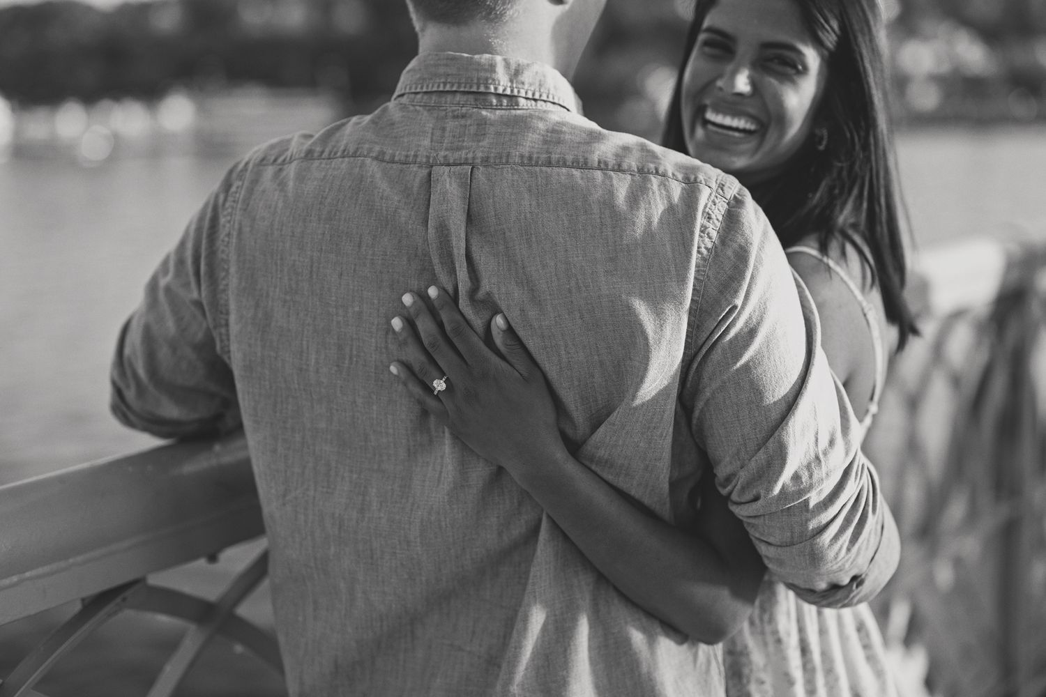 Black and white close-up of a woman smiling and holding a mans back near a waterfront.