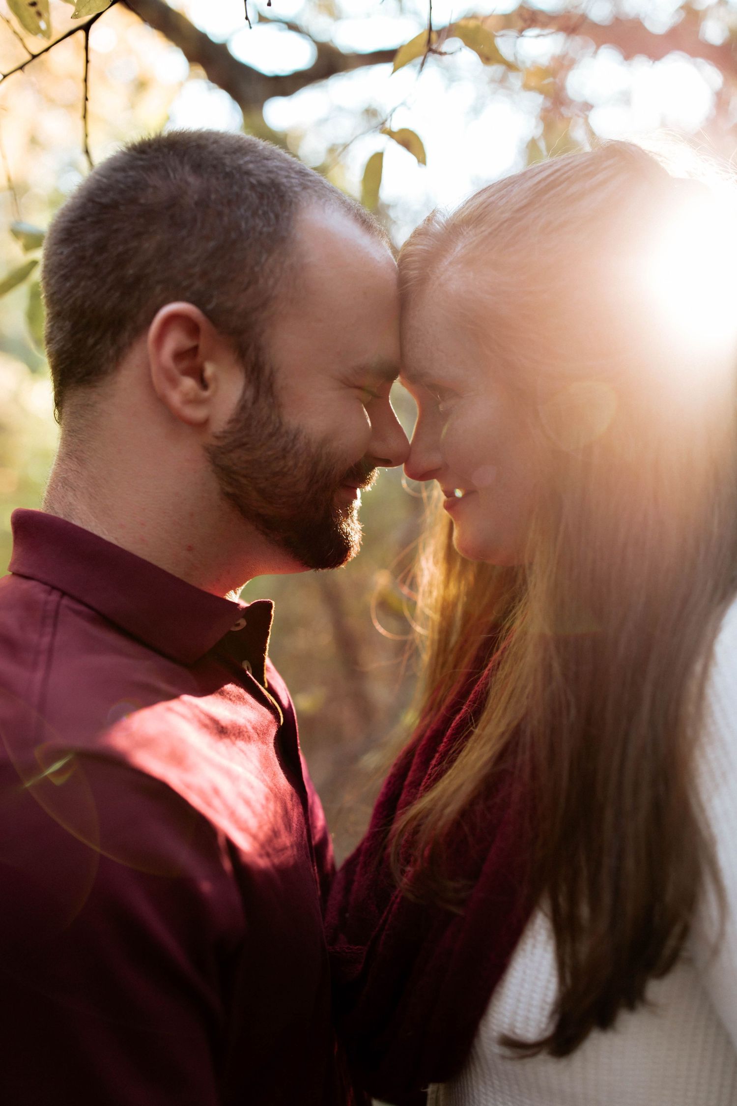 A bearded man and woman touch foreheads romantically in golden sunlight among autumn trees.