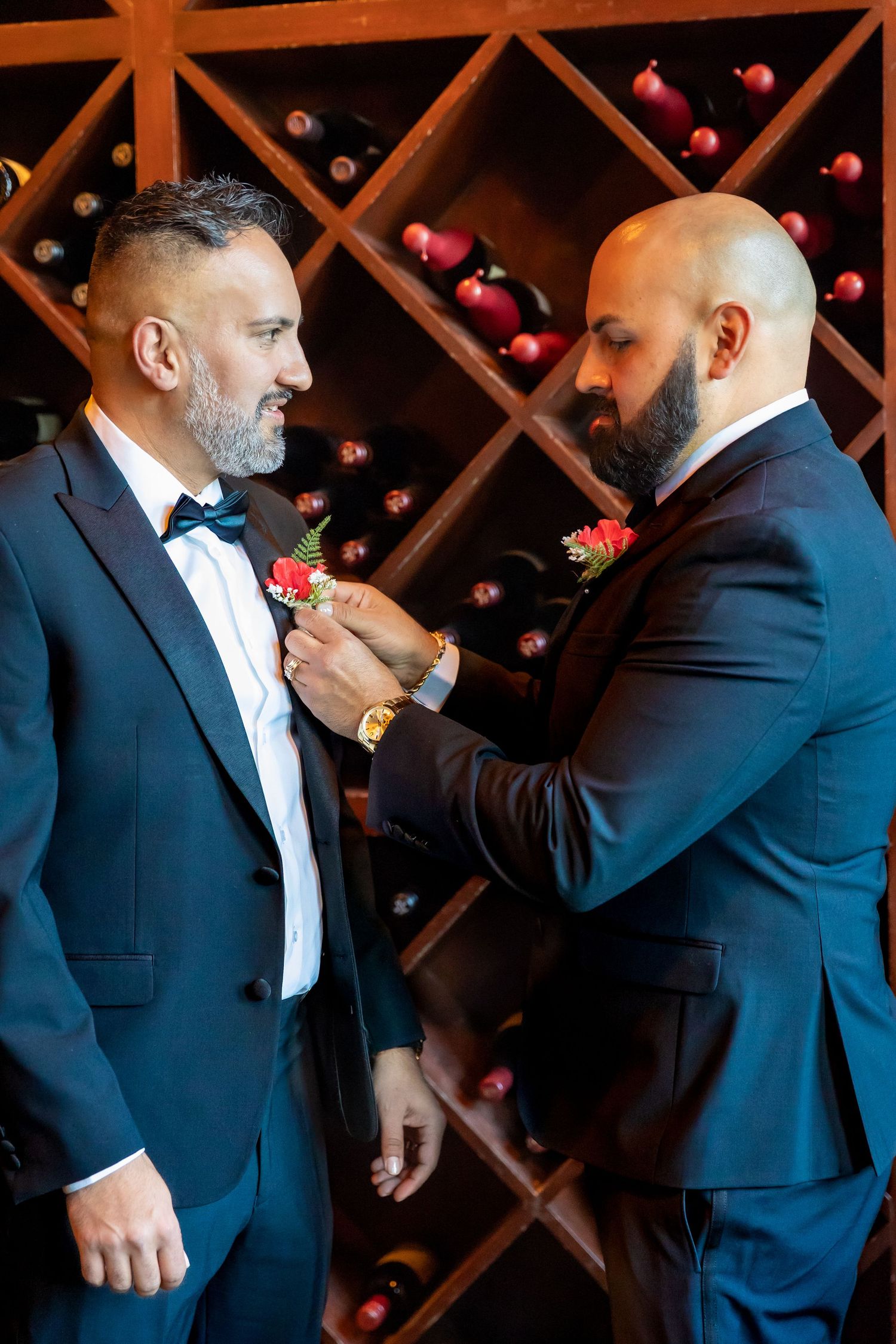 Two grooms in navy suits pin boutonnieres in a wine cellar during wedding preparations.