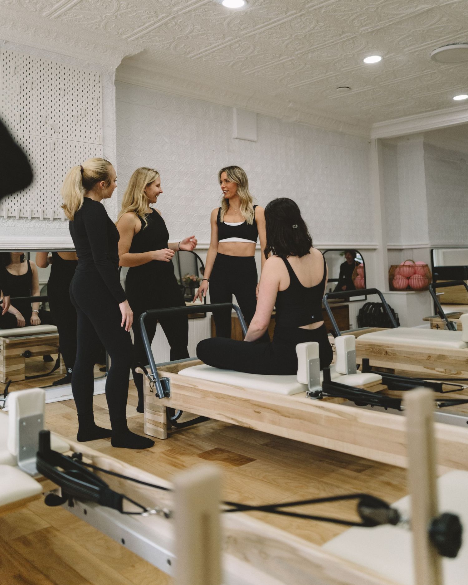 Women in black activewear socializing on Pilates reformer equipment in a bright, modern studio.