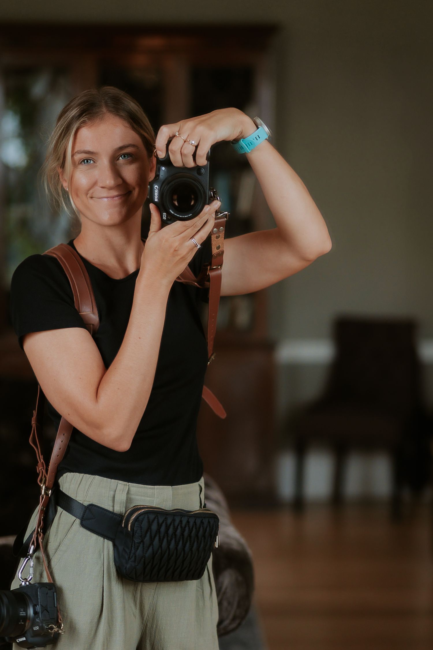 Smiling woman photographer holding a DSLR camera wearing a black t-shirt and green pants with a brown leather strap.