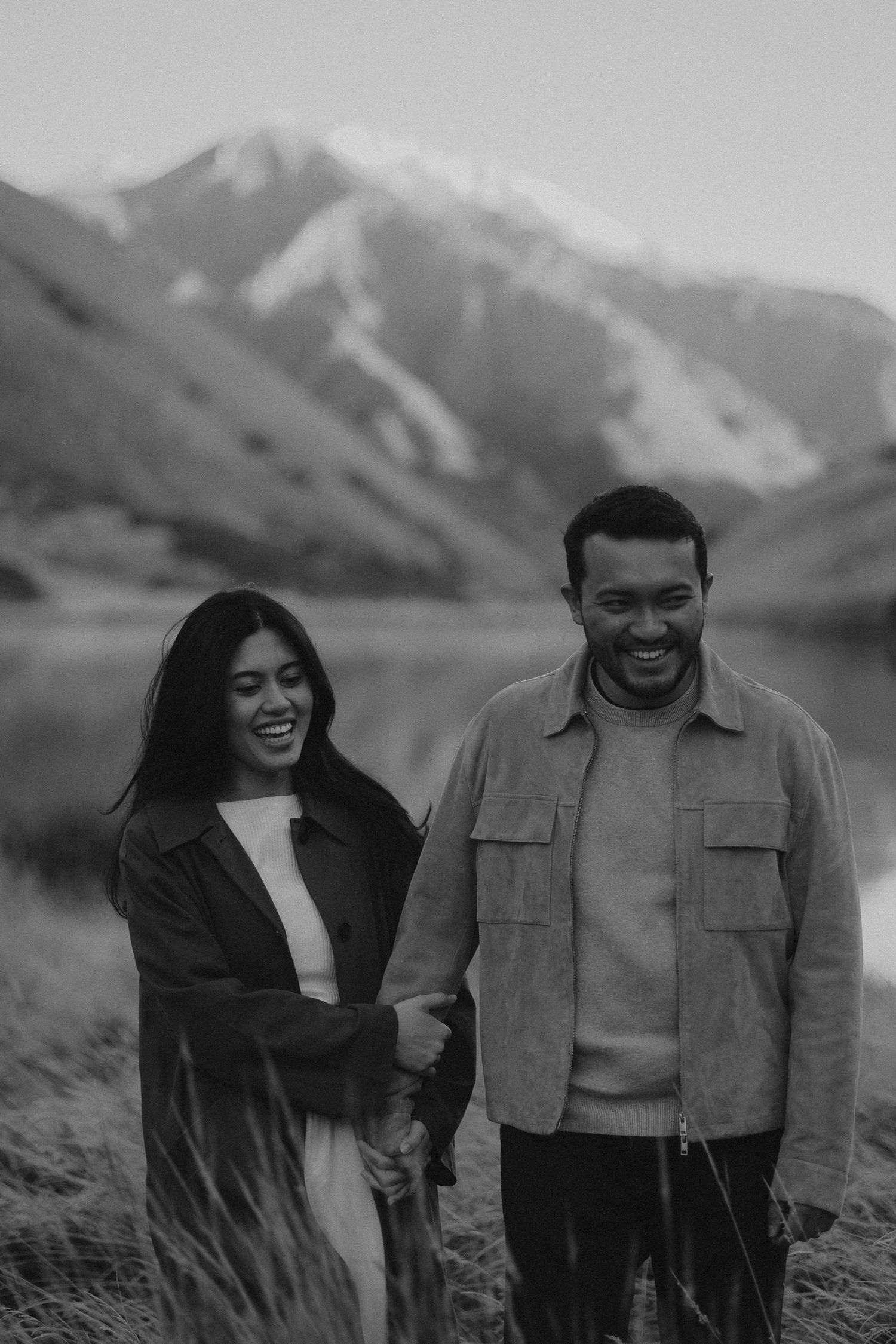 Black and white photo of a smiling couple holding hands with snow-capped mountains and a lake in the background.