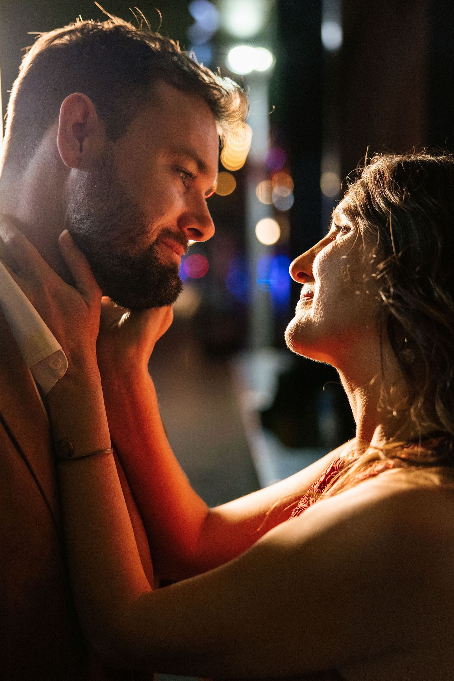 A romantic couple shares an intimate moment at night, with the woman tenderly holding the bearded mans face in city lights.