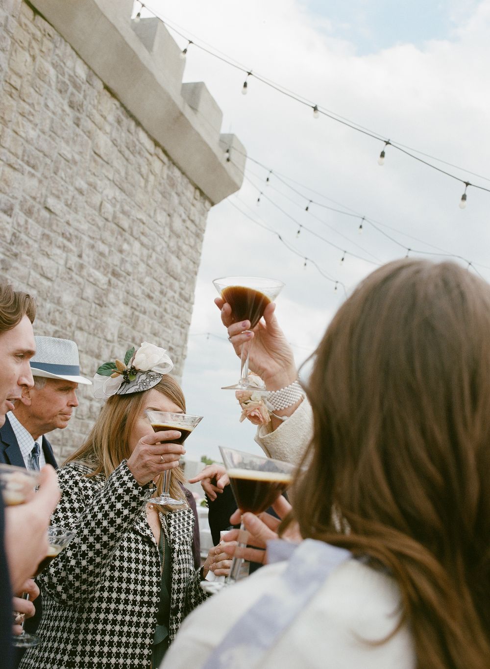 Wedding guests raising cocktails in a toast outdoors near a stone castle wall with string lights.