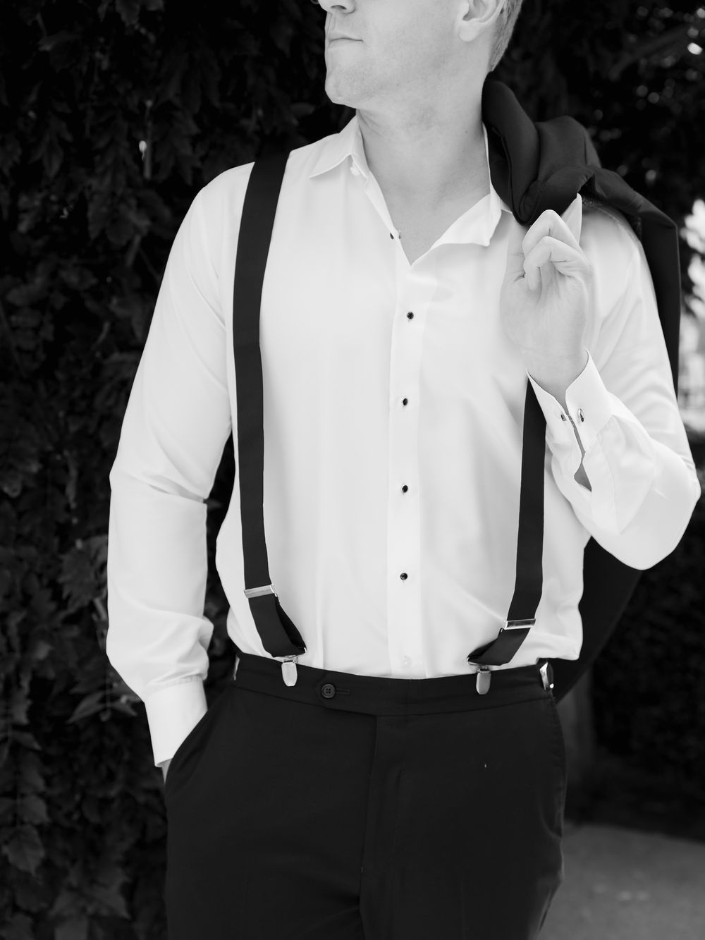 Black and white portrait of groom in white dress shirt with suspenders holding jacket over shoulder.