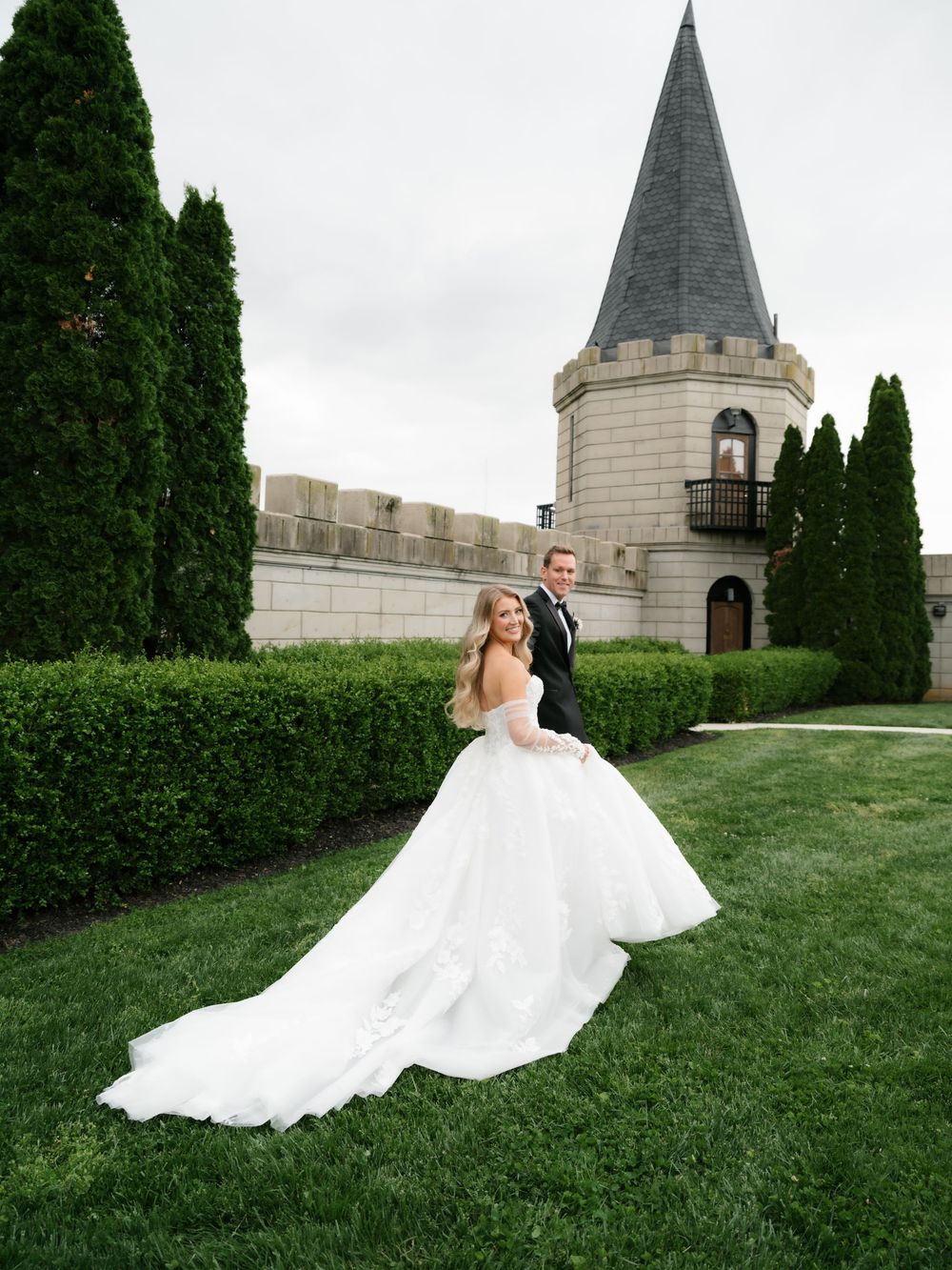 Bride in stunning white ball gown with long train poses with groom outside a castle venue on lush green grounds.