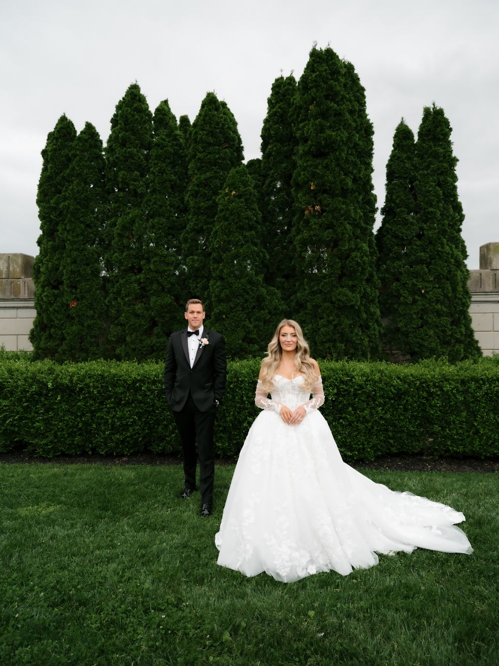 Bride in a white ball gown and groom in a black tuxedo pose on green grass with tall evergreen trees behind them.