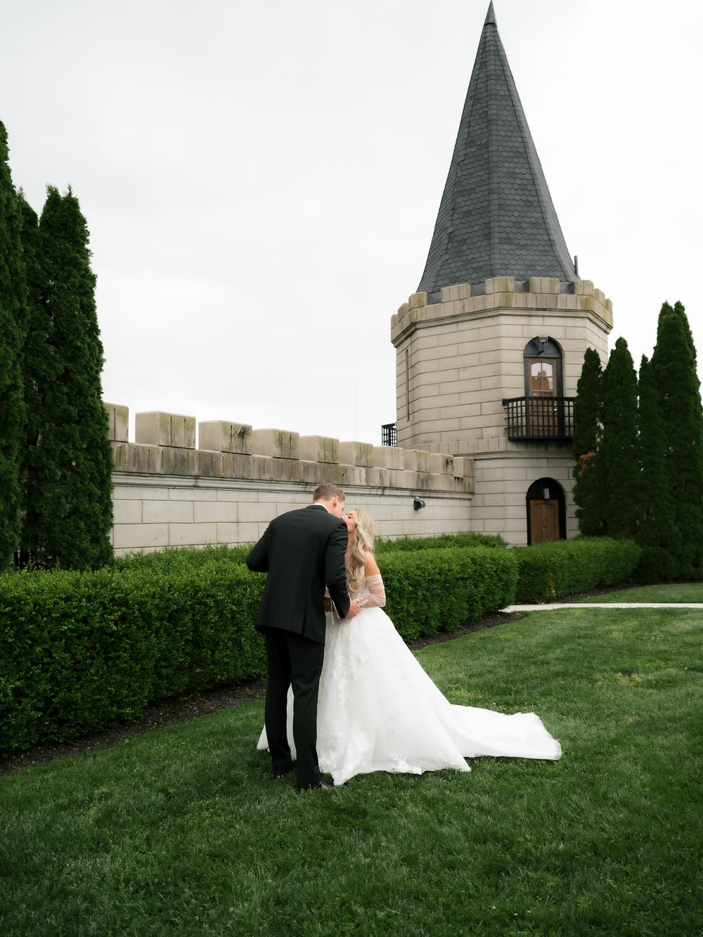 Bride and groom share romantic moment on lush green lawn beside castle-style wedding venue tower.