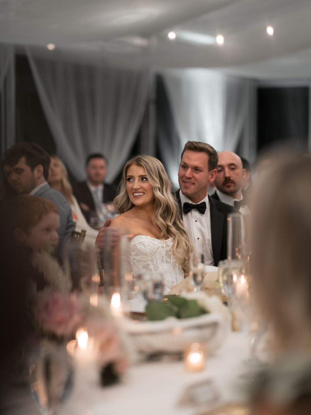 Bride in lace gown and groom in tuxedo sit at candlelit wedding reception head table surrounded by guests.