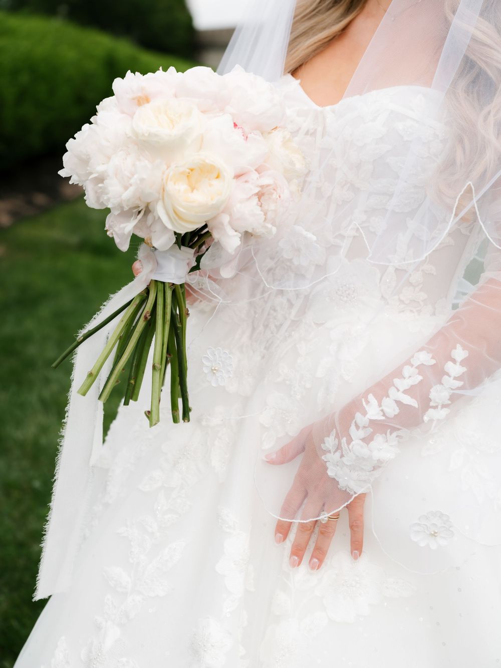 Bride holding a lush white and blush peony bouquet in a lace wedding gown with floral gloves.