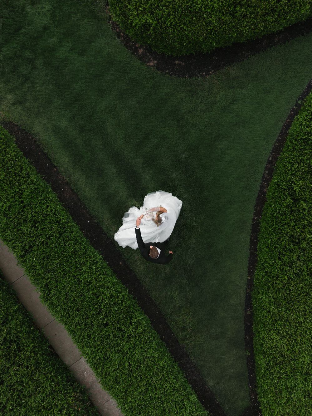 Aerial drone view of a bride and groom in white wedding dress lying on lush green manicured garden lawn.