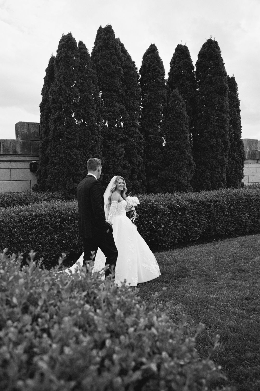 Bride and groom walking hand in hand through lush garden with tall evergreen trees in black and white wedding photo.