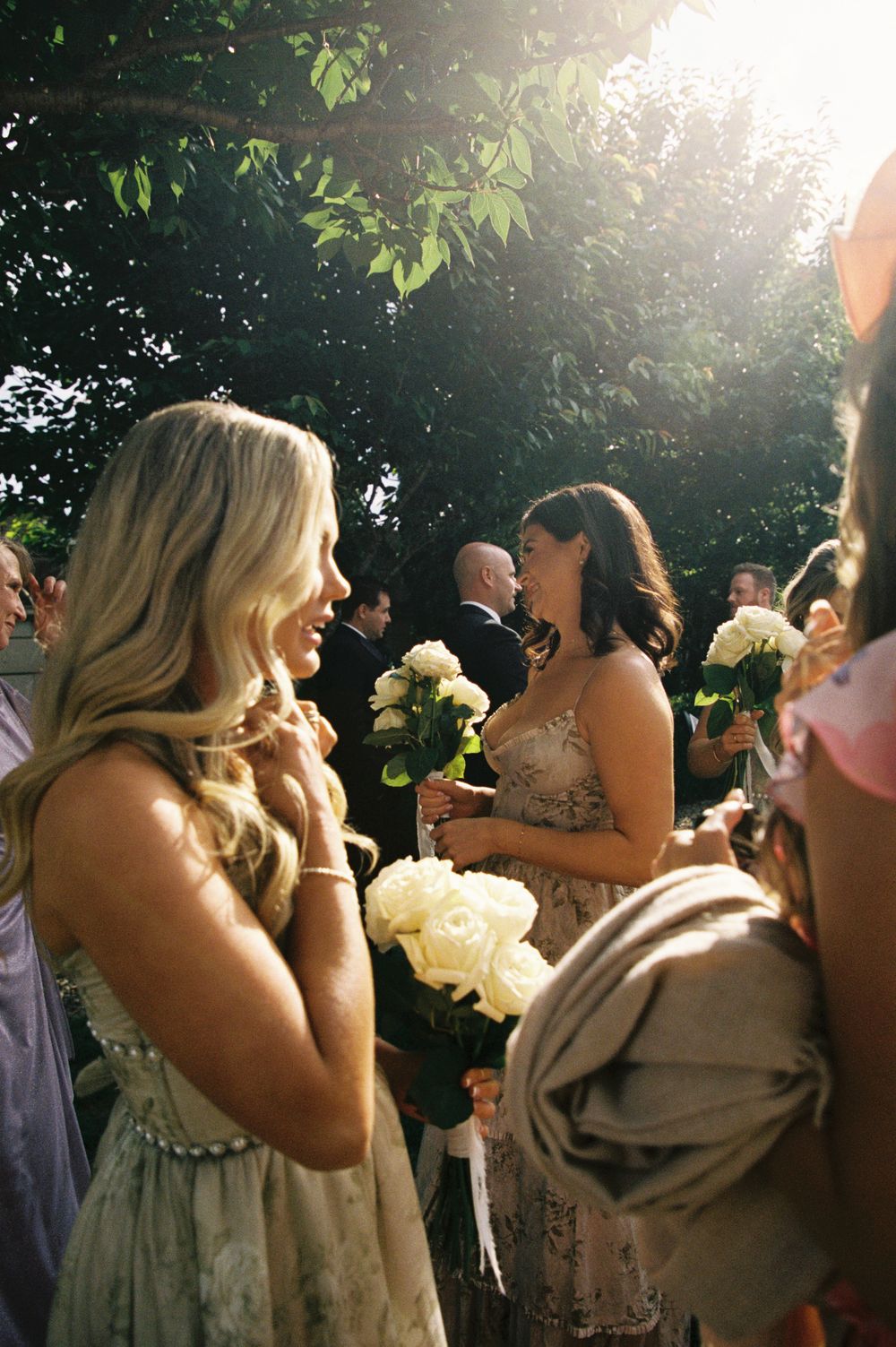 Bridesmaids holding white rose bouquets chatting in golden sunlight beneath a lush green tree.